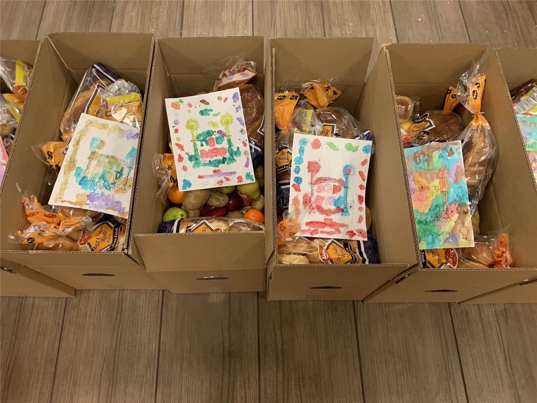 Packed Shabbos boxes with challah, fruit and vegetables, each topped with a card colored by a child